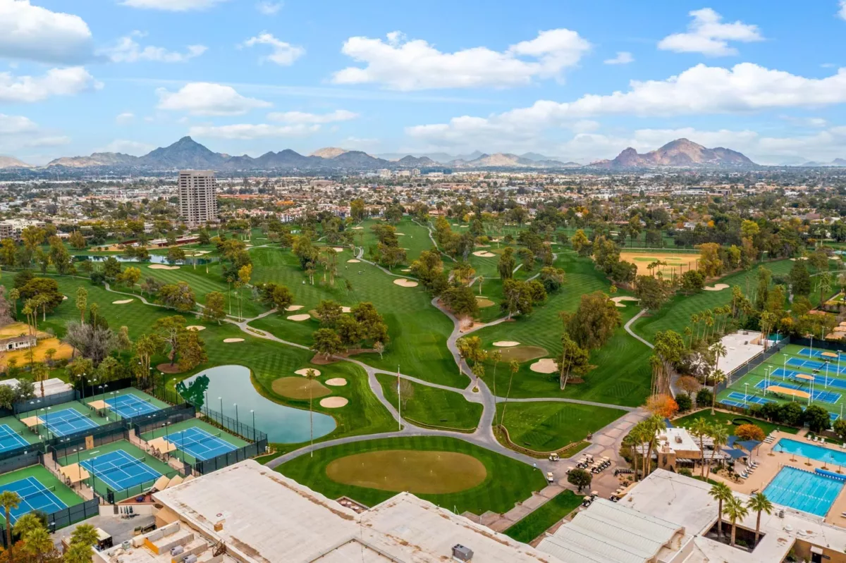Aerial view of golf course, tennis courts, and city skyline with mountain backdrop.